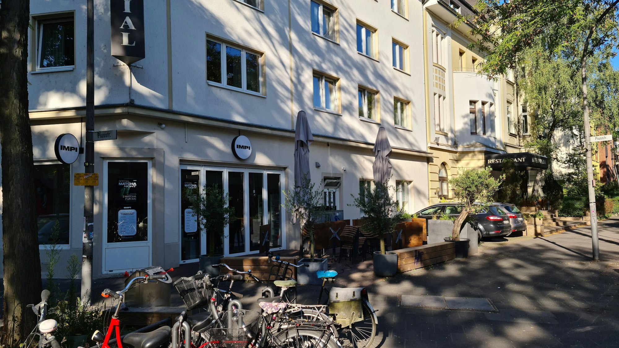 Viele Fahrräder stehen an Fahrradnadeln, dahinter ist ein Eck-Ladenlokal zu sehen, das Impi.