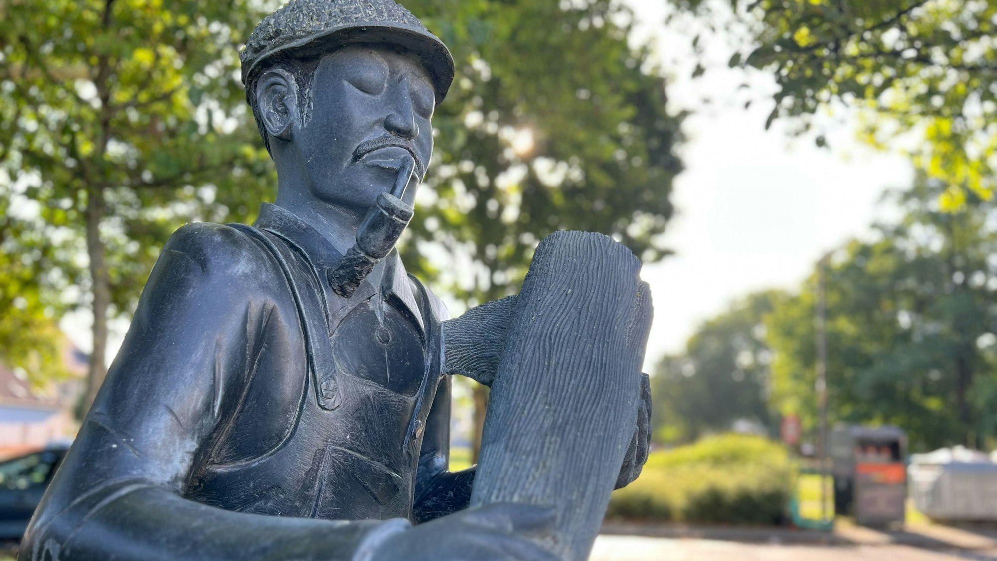 Die Skulptur eines Mannes, der einen Besen bindet, ist zu sehen. Er sitzt draußen auf einem Platz inmitten einer Rasenfläche.