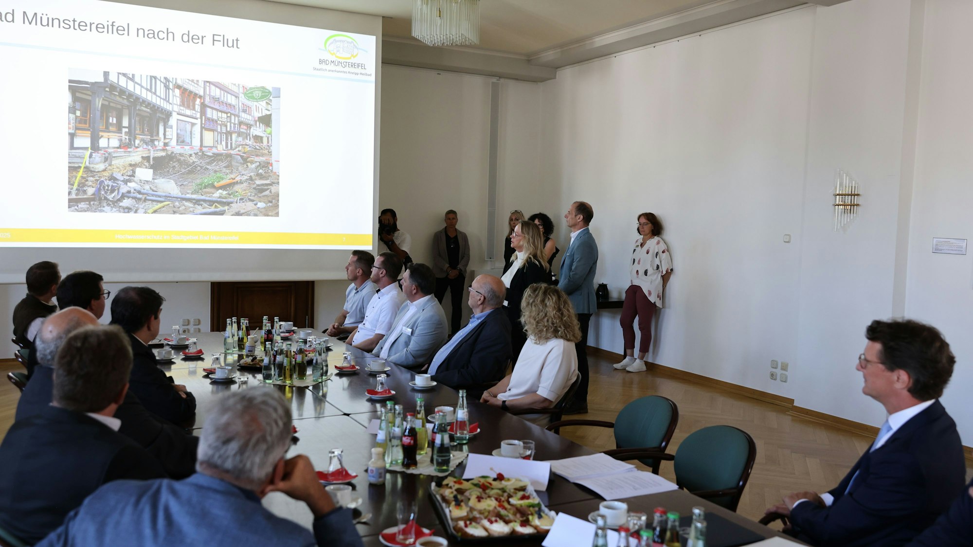 Auf einer Leinwand ist die nach der Flut mit Trümmern übersäte Innenstadt von Bad Münstereifel zu sehen. Die Besucher, auch der Ministerpräsident, verfolgen die Ausführungen der Bürgermeisterin.