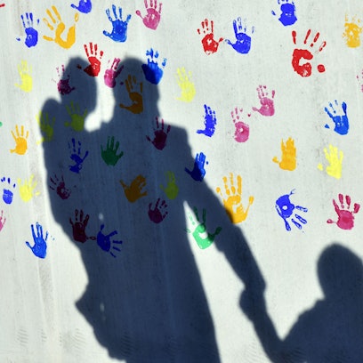 Ein Mann mit einem Kind auf dem Arm und einem an der Hand wirft einen Schatten auf eine mit bunten Handabdrücken bemalte Wand einer Kindertagesstätte.