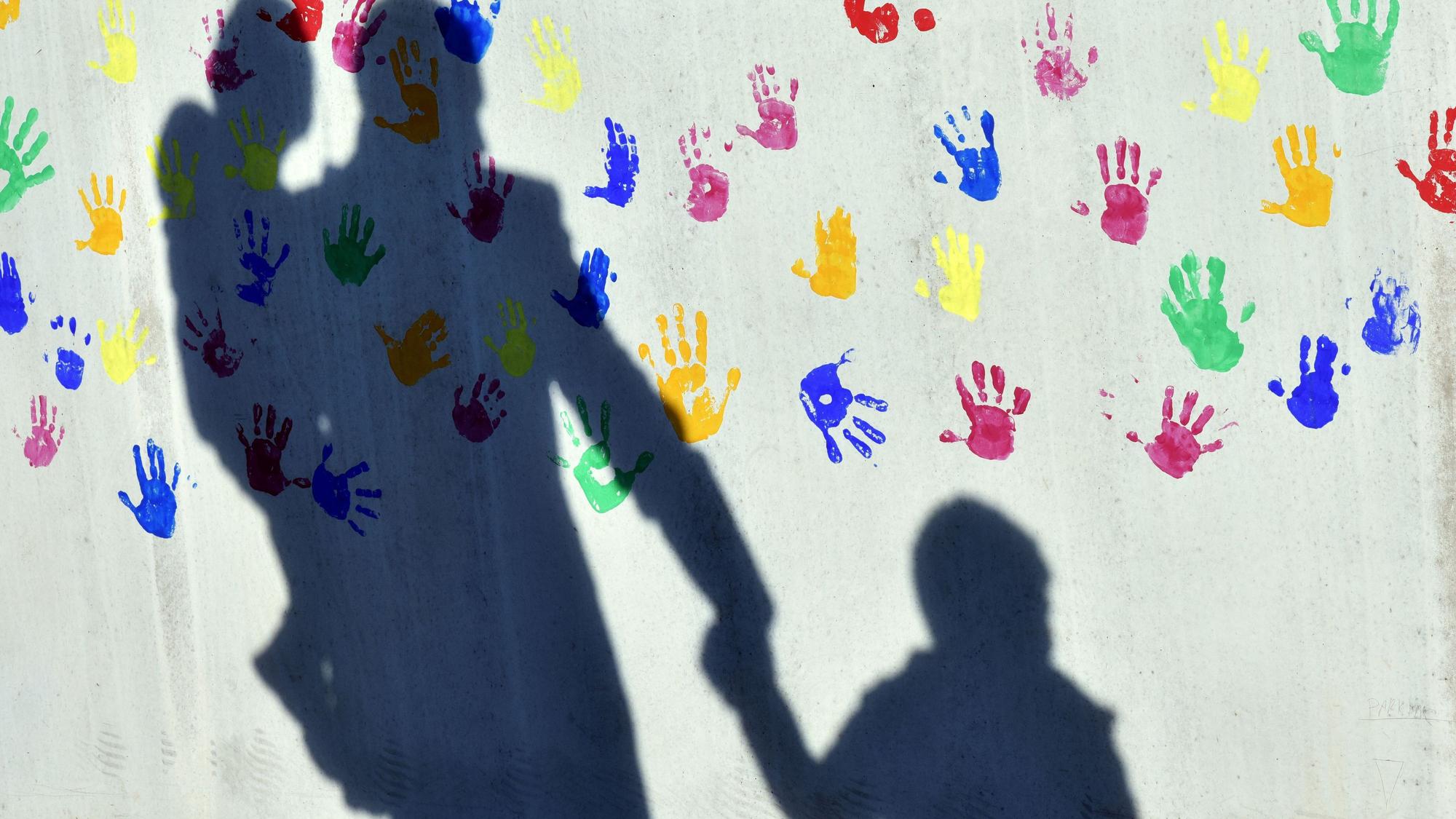 Ein Mann mit einem Kind auf dem Arm und einem an der Hand wirft einen Schatten auf eine mit bunten Handabdrücken bemalte Wand einer Kindertagesstätte.