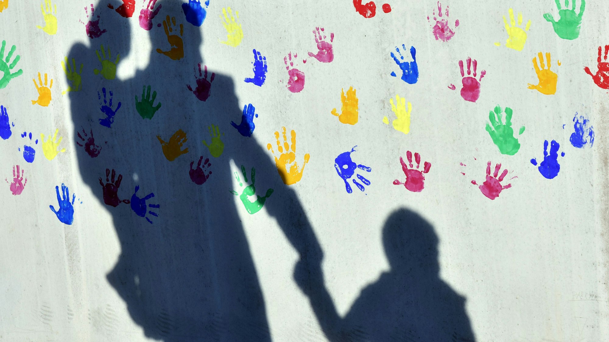 Ein Mann mit einem Kind auf dem Arm und einem an der Hand wirft einen Schatten auf eine mit bunten Handabdrücken bemalte Wand einer Kindertagesstätte.