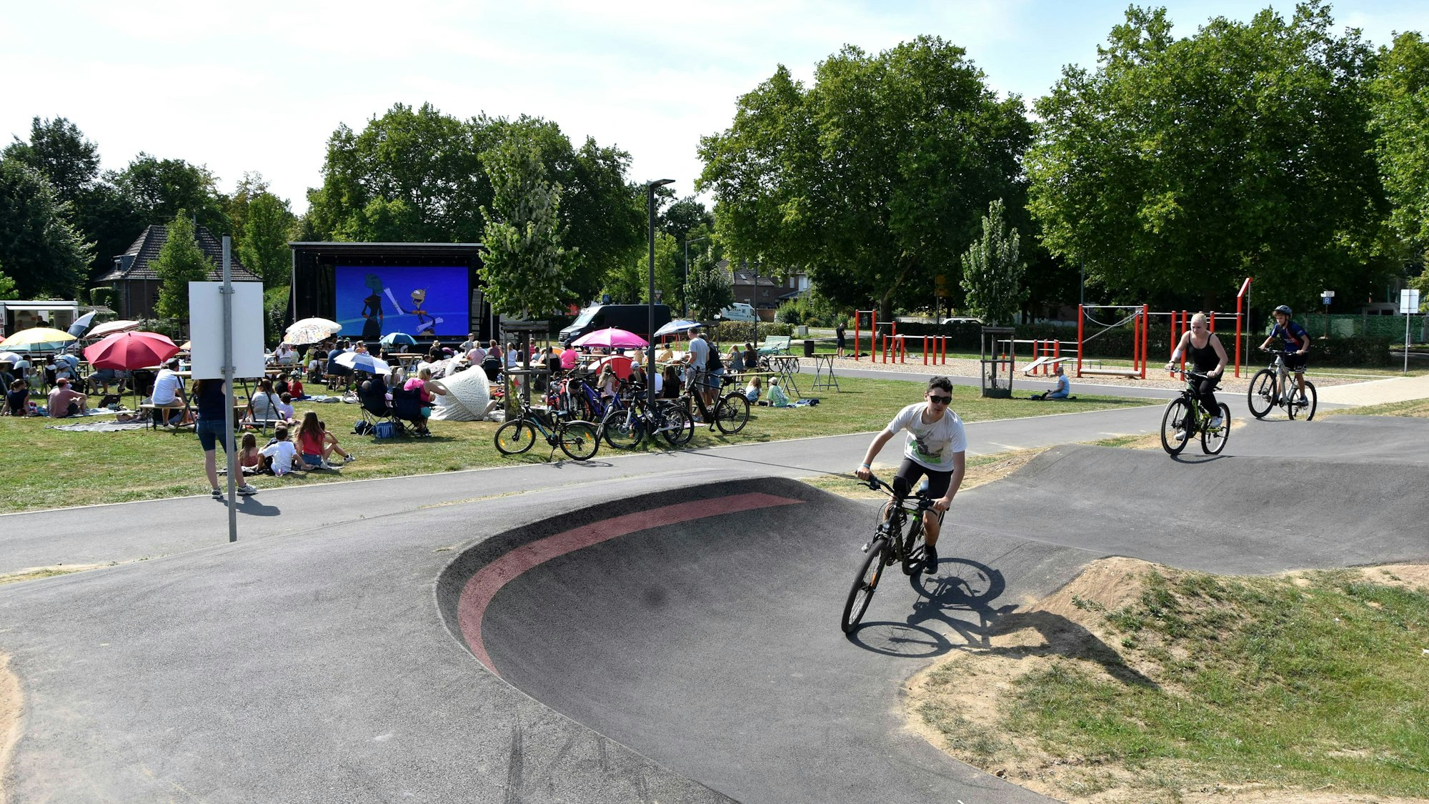 Jugendliche fahren auf ihren Mountainbikes auf der Strecke, im Hintergrund haben viele auf einer Wiese Platz genommen, um auf einer großen Leinwand den Film zu sehen