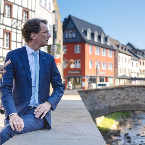 Auf der Erftmauer sitzend blickt Hendrik Wüst auf die andere Seite der Erft, während Sabine Preiser-Marian gestikulierend erklärt. Im Hintergrund die Aktstadtkulisse von Bad Münstereifel.