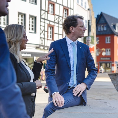 Auf der Erftmauer sitzend blickt Hendrik Wüst auf die andere Seite der Erft, während Sabine Preiser-Marian gestikulierend erklärt. Im Hintergrund die Aktstadtkulisse von Bad Münstereifel.