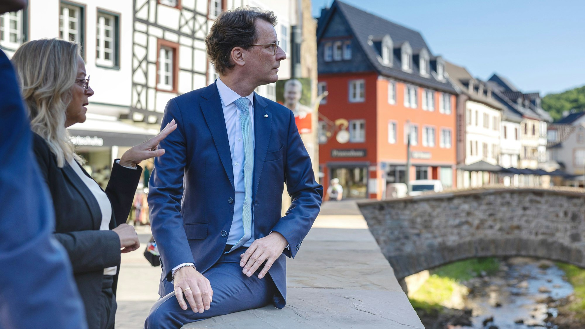 Auf der Erftmauer sitzend blickt Hendrik Wüst auf die andere Seite der Erft, während Sabine Preiser-Marian gestikulierend erklärt. Im Hintergrund die Aktstadtkulisse von Bad Münstereifel.