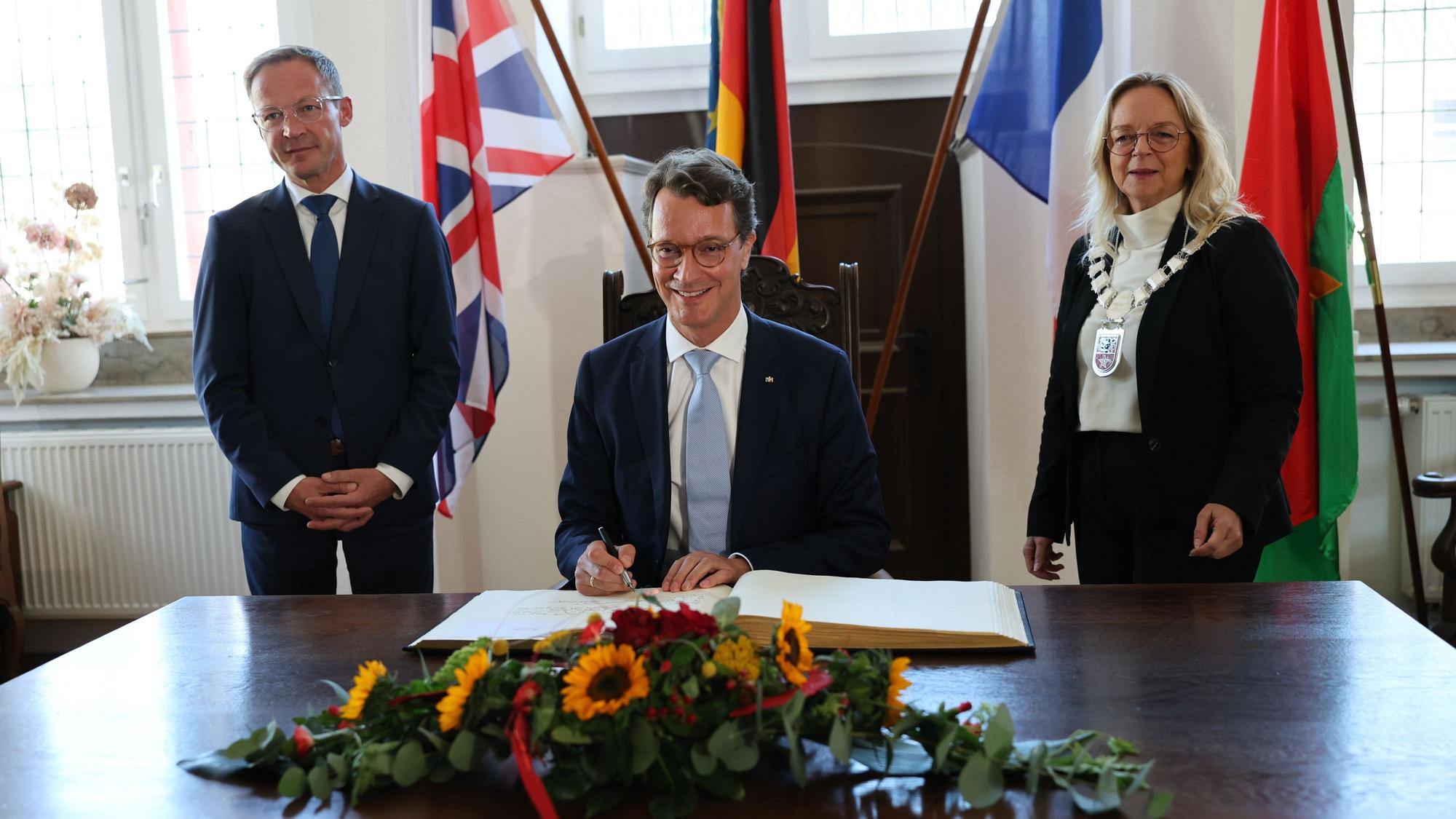 Ministerpräsident Hendrik Wüst schreibt ins Goldene Buch, neben ihm die Bürgermeisterin und der Staatssekretär. Im Hintergrund sind Fahnen zu sehen.