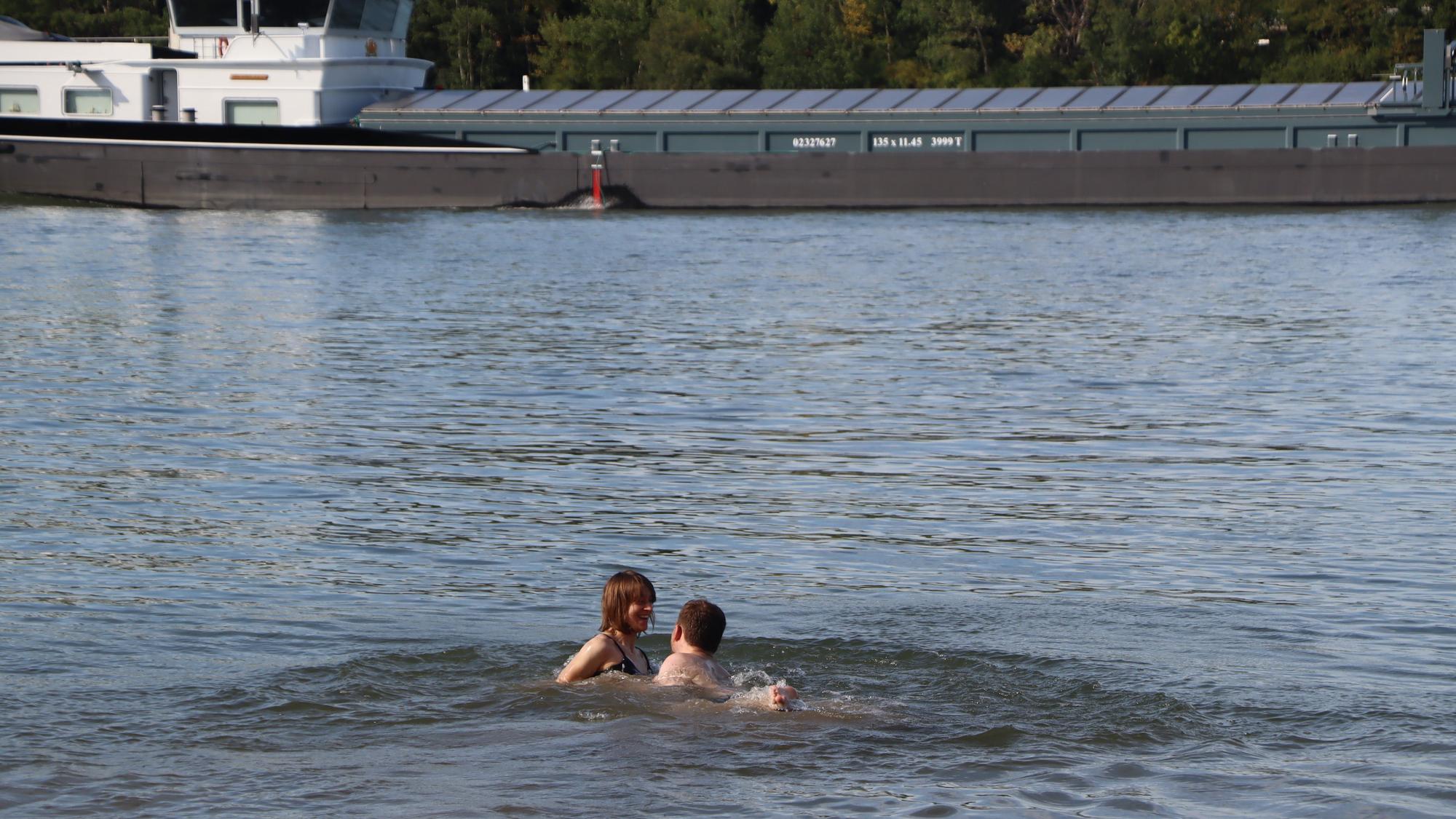 Zwei Personen schwimmen im Rhein, in Hintergrund ein Frachtschiff.