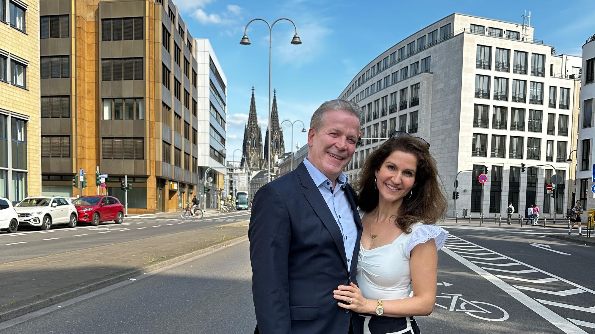 Auf der Gereonstraße stehen ein Mann und eine Frau und lächeln in die Kamera. Im Hintergrund ist der Dom zu sehen.