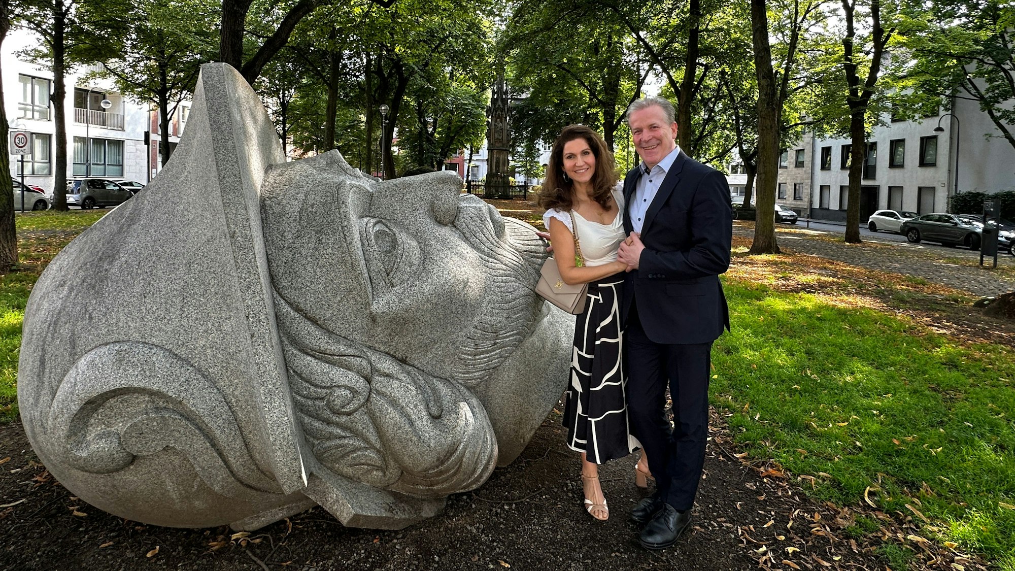 Eine Skulptur im Gereonsdriesch mit Mariensäule. Daneben stehen eine Frau und ein Mann und lächeln in die Kamera.