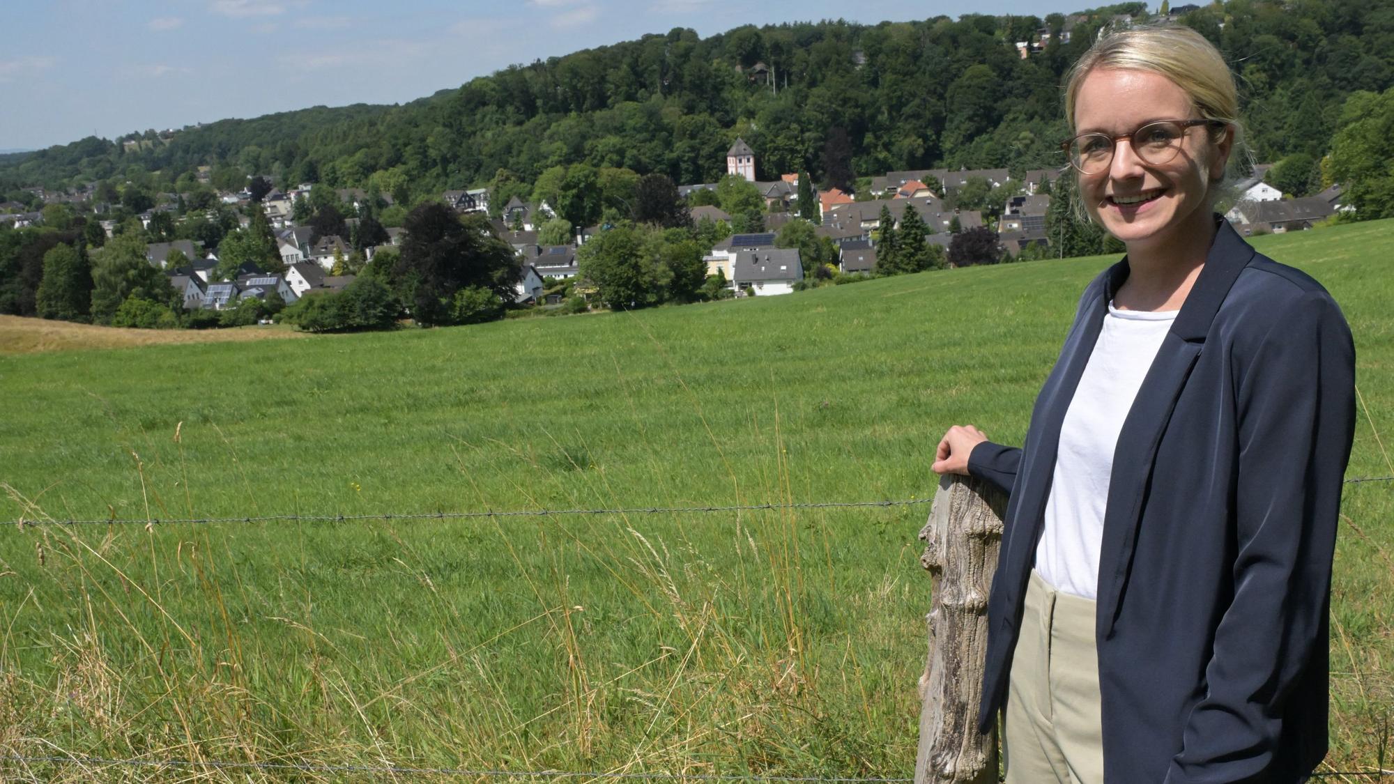 Eine junge Frau steht vor einer Wiese, unten im Tal ein kleines Örtchen mit Kirche.