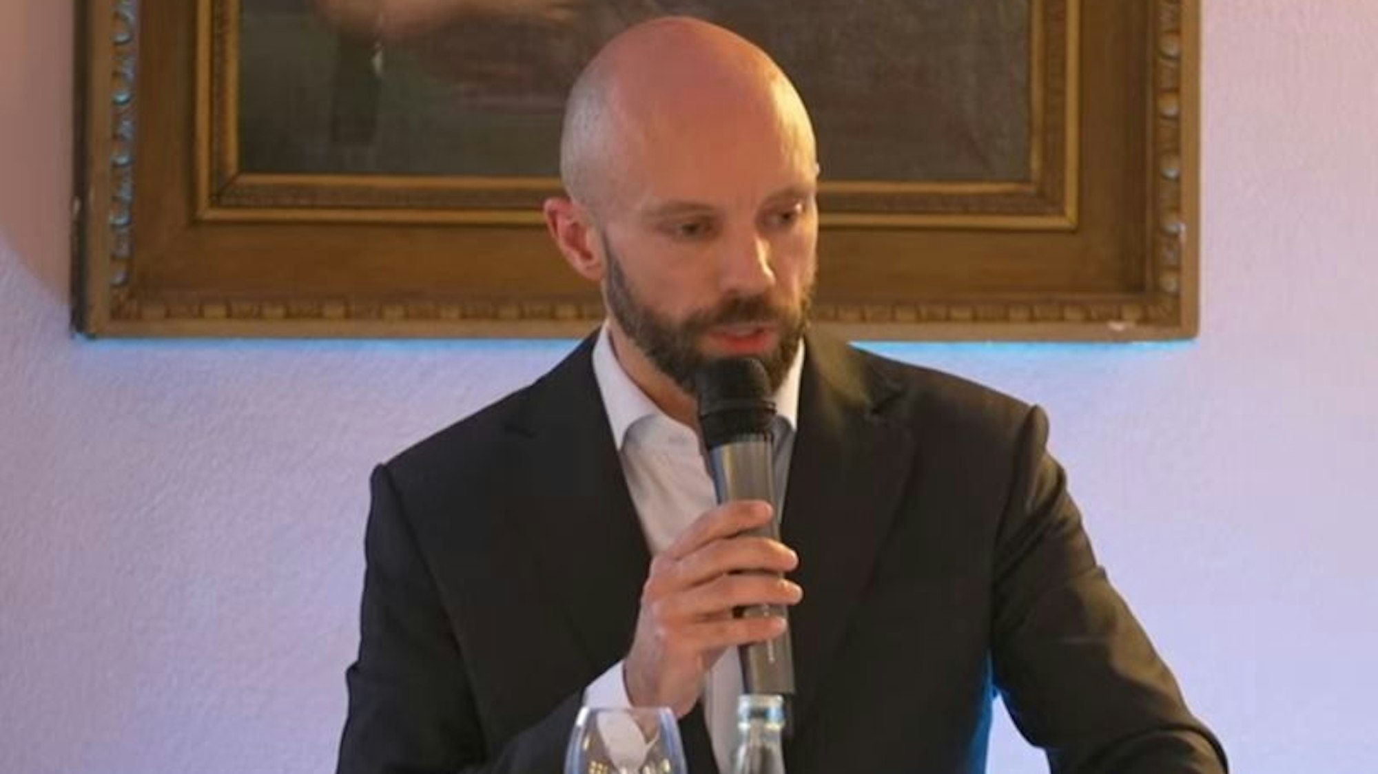 Auf dem Foto ist ein Mann mit Glatze und Vollbart zu sehen. Er hält ein Mikrofon in der Hand.