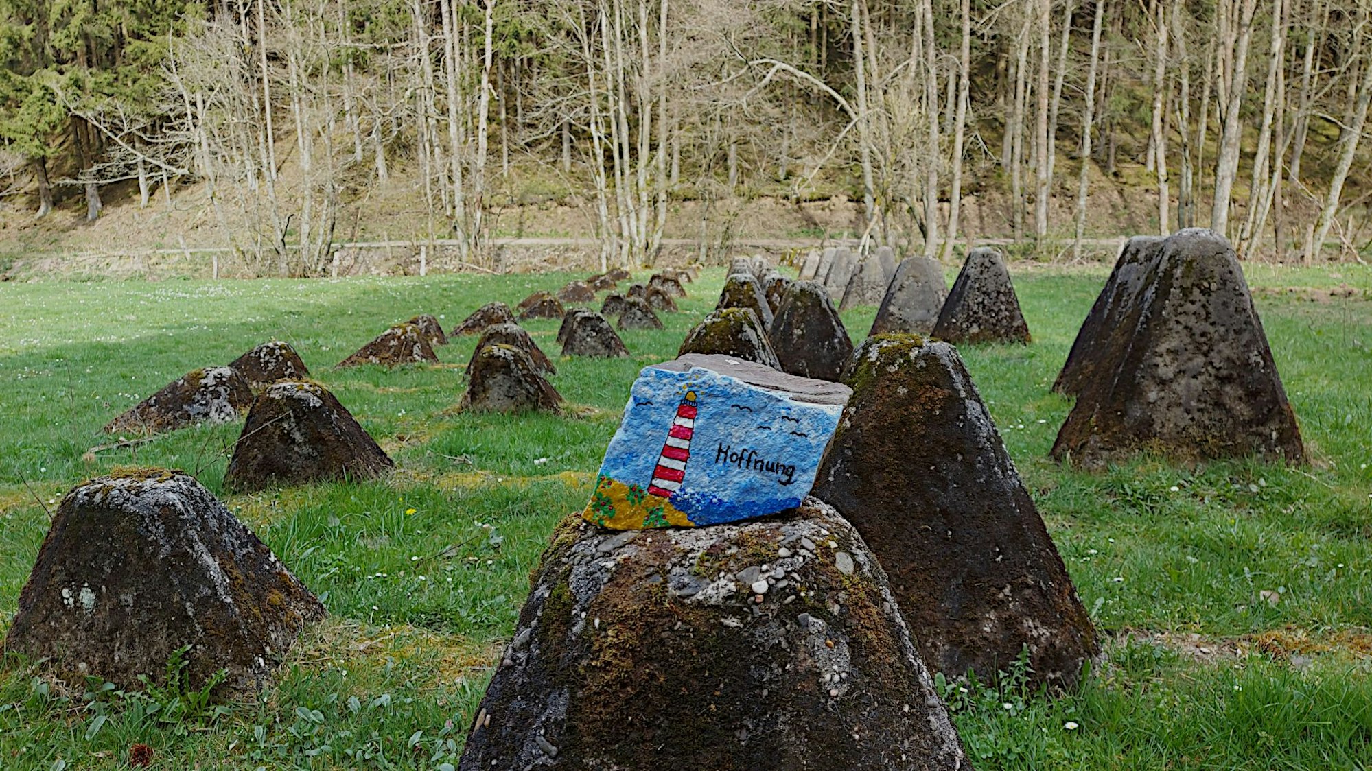Auf eine Wiese vor einem Waldrand verläuft eine Reihe von Panzersperren. Auf einem der Betonklötze liegt ein bunt bemalter Stein.