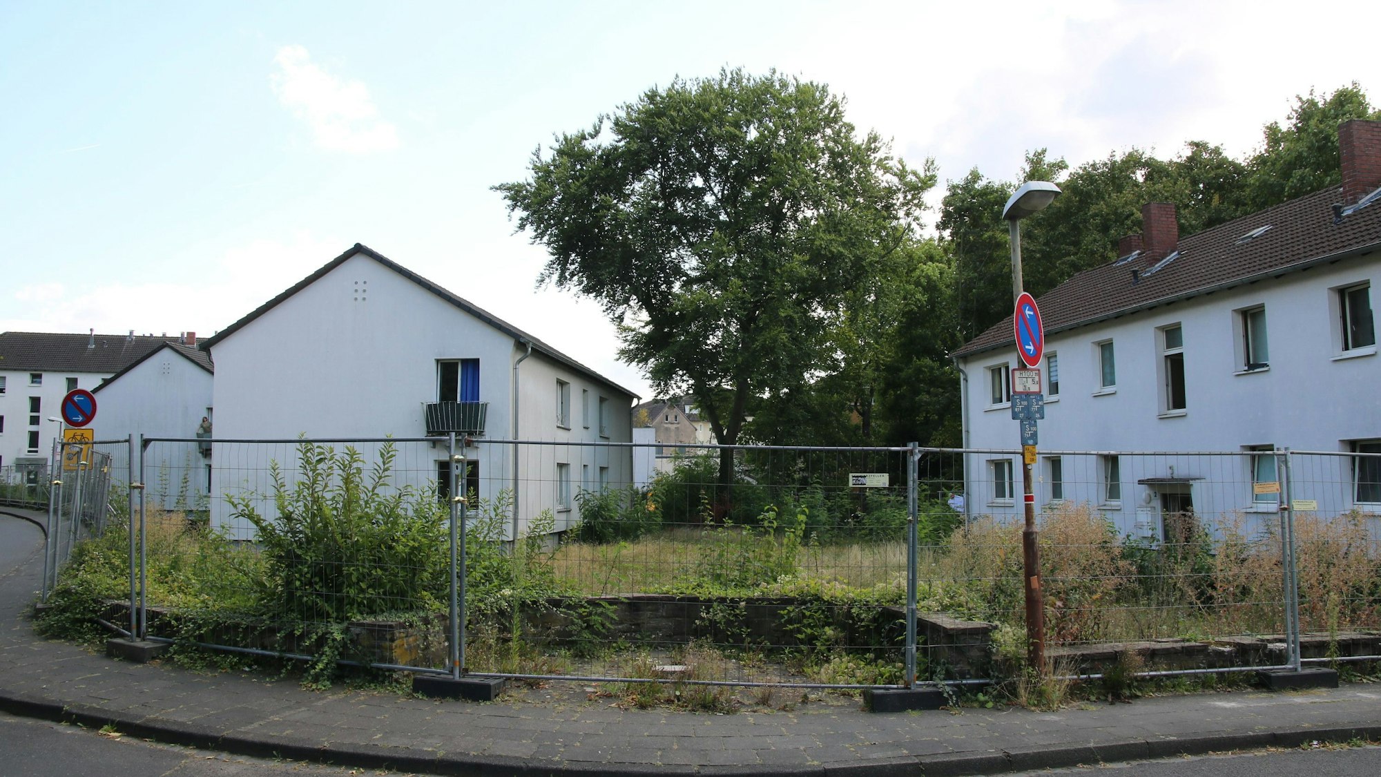 Drei zweigeschossige Gebäude mit einer Grünfläche, alles ist umgeben mit einem Bauzaun.