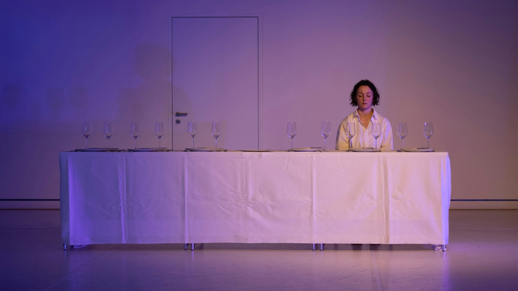 Eine Frau sitzt an einer langen, mit einer weißen Tischdecke gedeckten Tafel. Auf dem Tisch stehen 12 Weingläser.