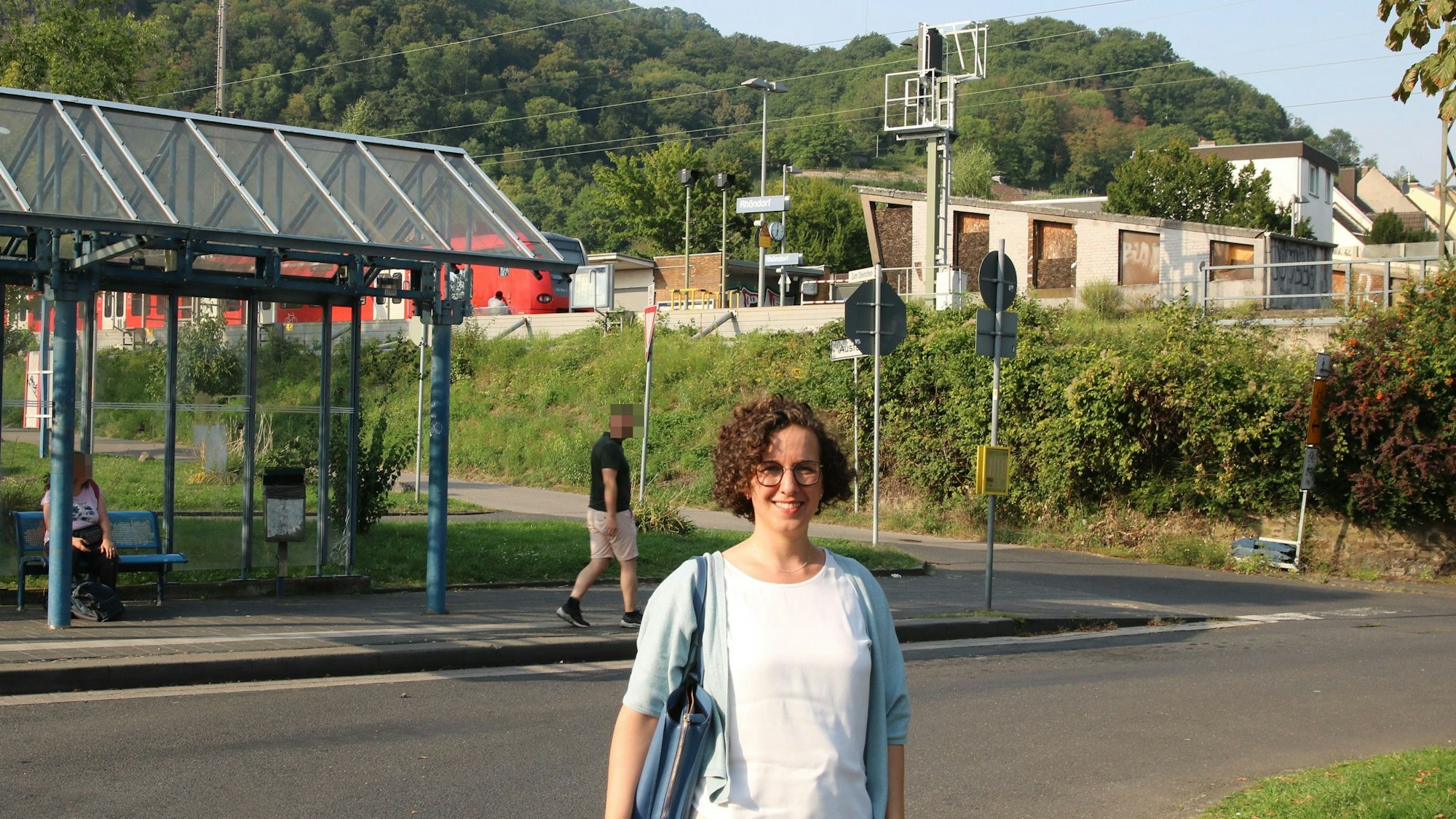 Eine Frau an einer Bushaltestelle, im Hintergrund der Bahnhof Rhöndorf-