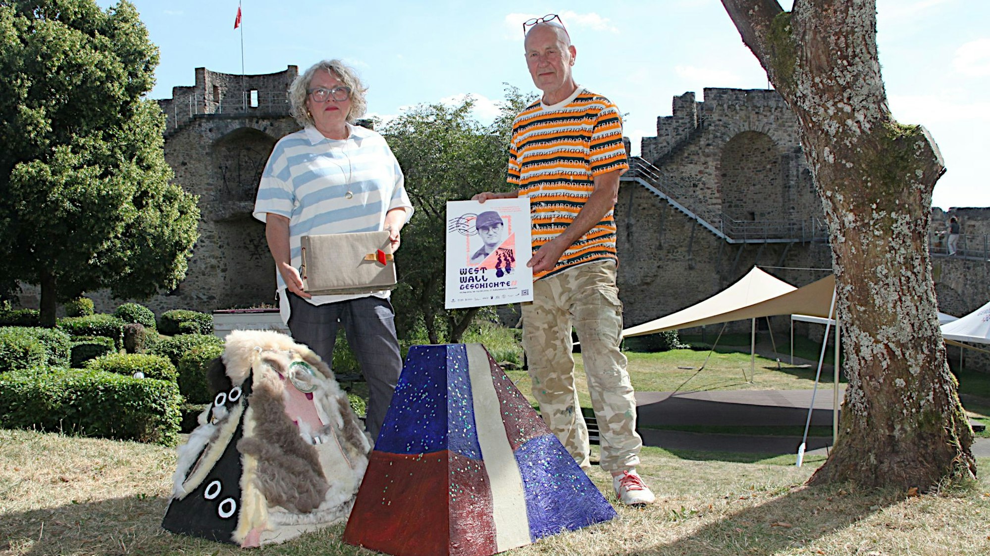 Claudia Warda und Jan Pronk stehen hinter zwei nachgebauten „Höckern“. In den Händen halten sie Informationsmaterial zu dem Kunstprojekt.