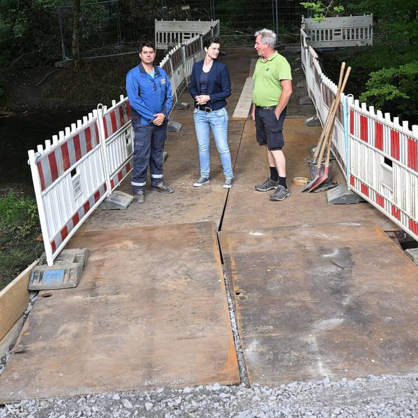 Zwei Männer und eine Frau stehen auf einer Behelfsbrücke aus Stahlplatten, die über einen Bach im Wald führt.