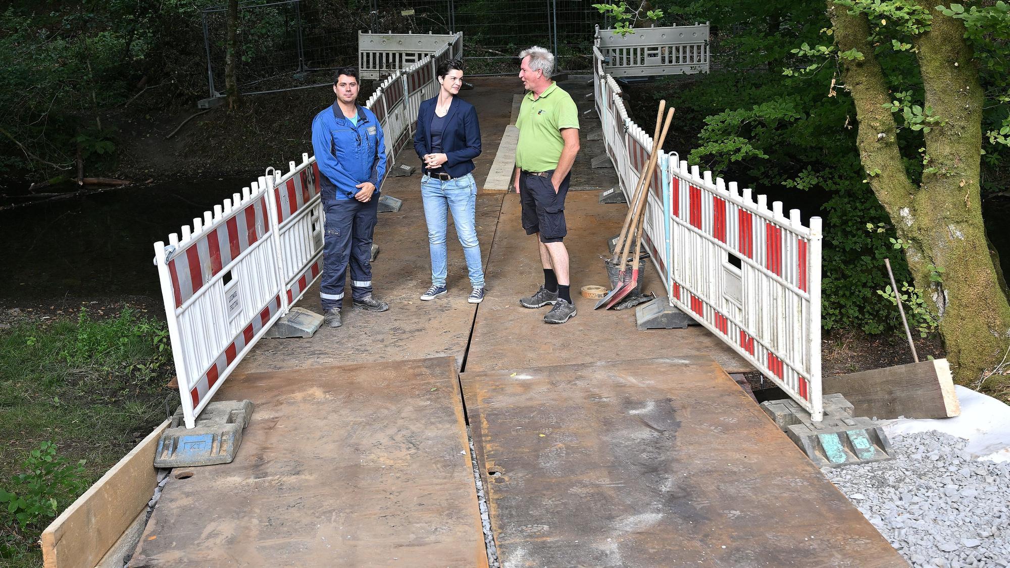 Zwei Männer und eine Frau stehen auf einer Behelfsbrücke aus Stahlplatten, die über einen Bach im Wald führt.
