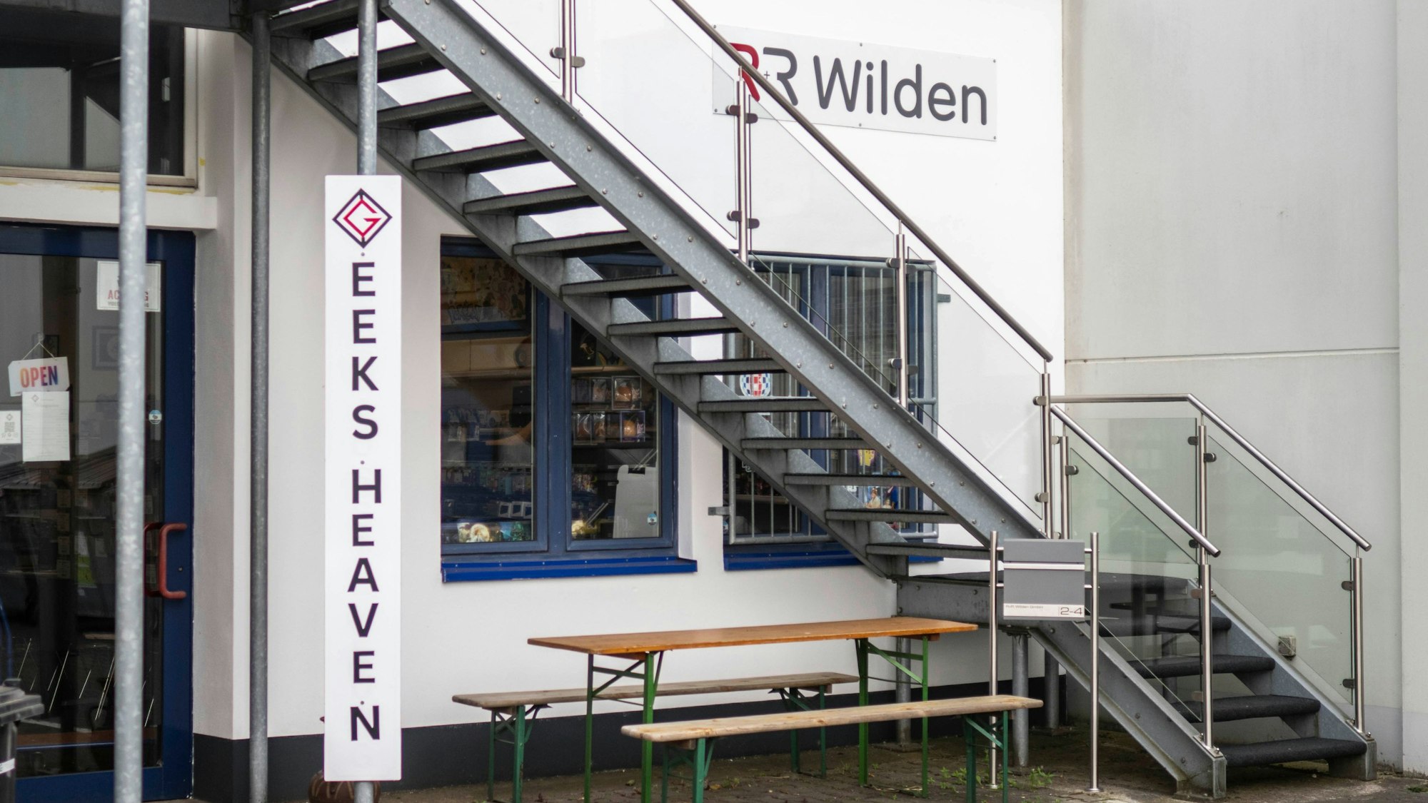 An einem Gebäude ist das Schild „Geeks Heaven“ angebracht. Ein Tisch und zwei Bänke stehen davor. Eine Stahltreppe für in die obere Etage.