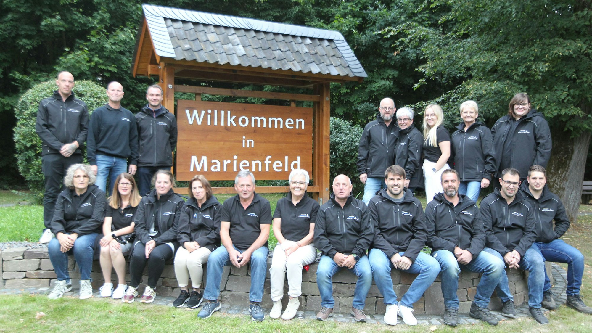 Eine Gruppe von Frauen und Männern sitzt und steht neben und vor einem hölzernen Schild mit der Aufschrift „Willkommen in Marienfeld“.