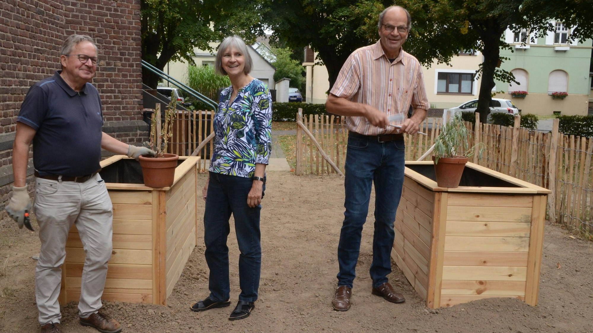 Zwei Männer und eine Frau stehen auf einem vorbereiteten Gartengrundstück. Der Boden ist kahl, zwei Hochbeete sind aufgebaut, aber noch nicht bepflanzt.