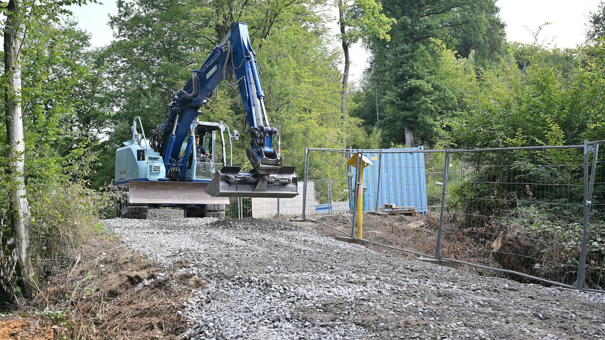 Ein blauer Bagger steht an einer Baustellenzufahrt im Wald, der Weg ist geschottert.