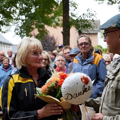 Christel Kastl steht links und hält einen Blumenstrauß und einen kleinen Ballon mit der Aufschrift „Danke“ in den Händen. Um sie herum stehen zahlreiche Menschen.