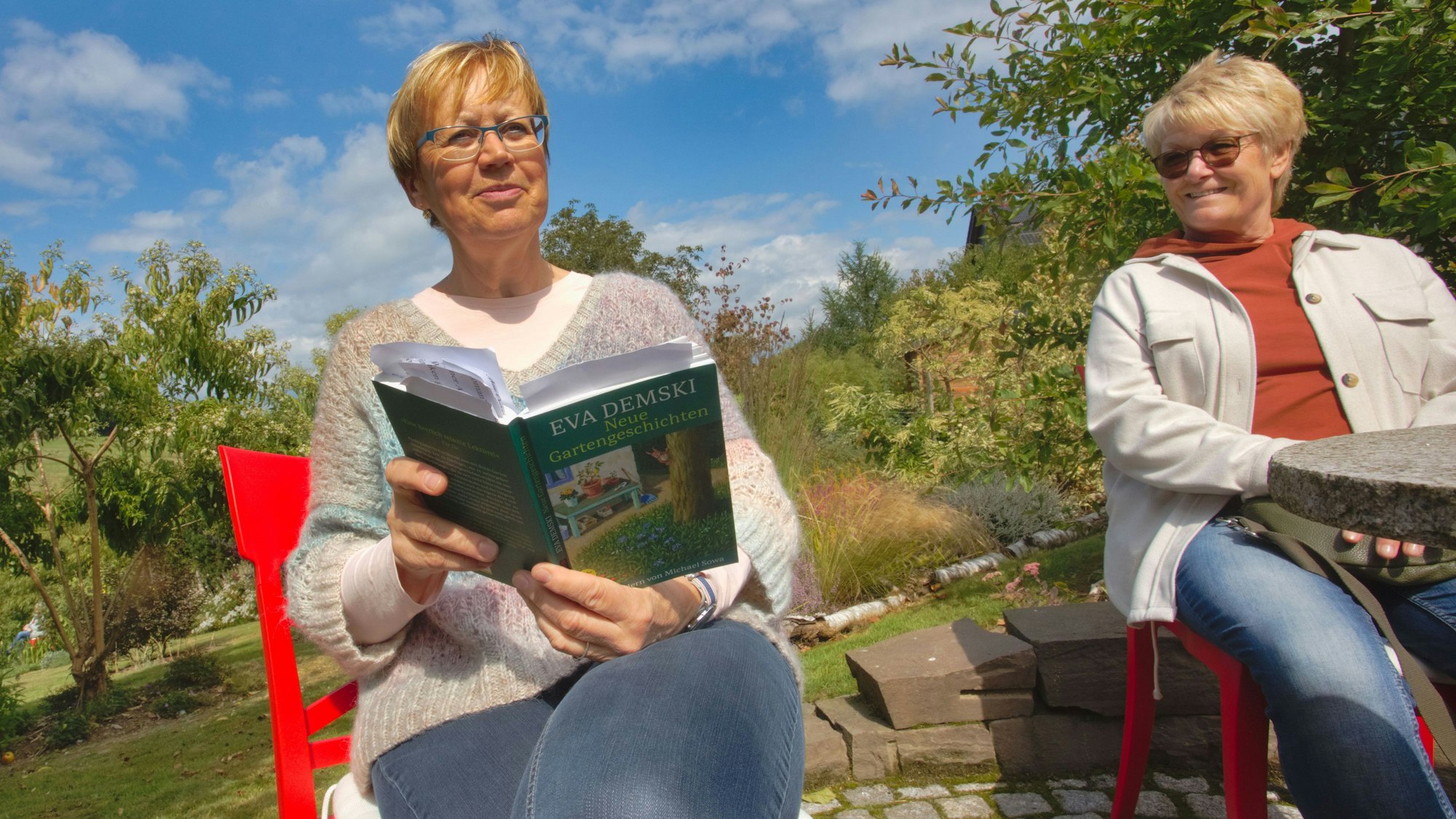 Das Foto zeigt zwei Frauen, die im Garzen sitzen, eine liest aus einem Buch vor