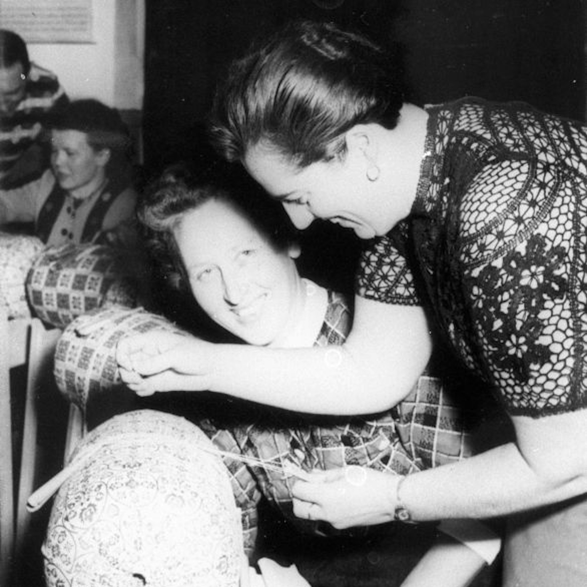 Eine Schwarz-weiß-Aufnahme zeigt sei Frauen, die sich angrinsen. Sie werden angeleuchtet. Die Frau im Vordergrund klöppelt ein Band.