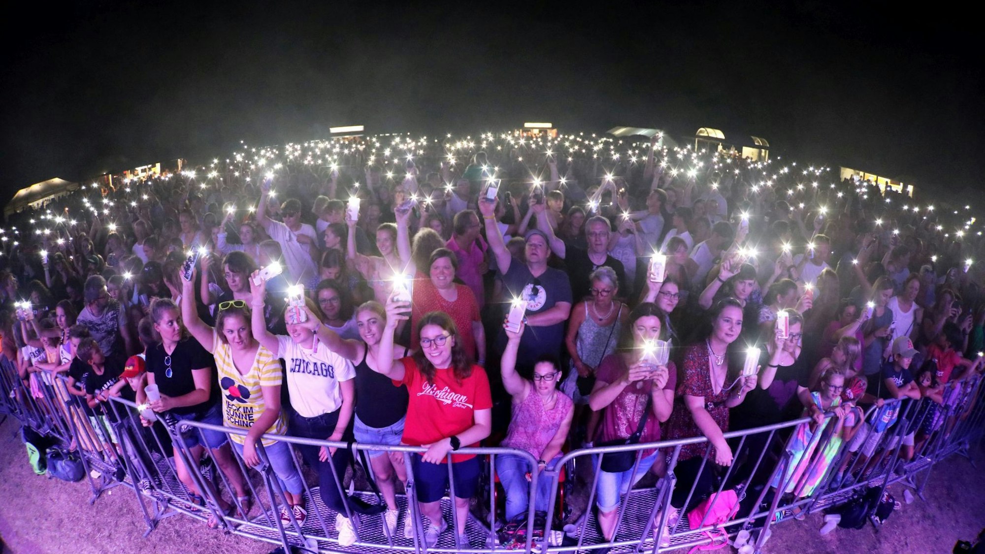 Hunderte Menschen stehen hinter einer Absperrung vor einer Bühne, viele leuchten mit den Taschenlampen ihrer Handys.