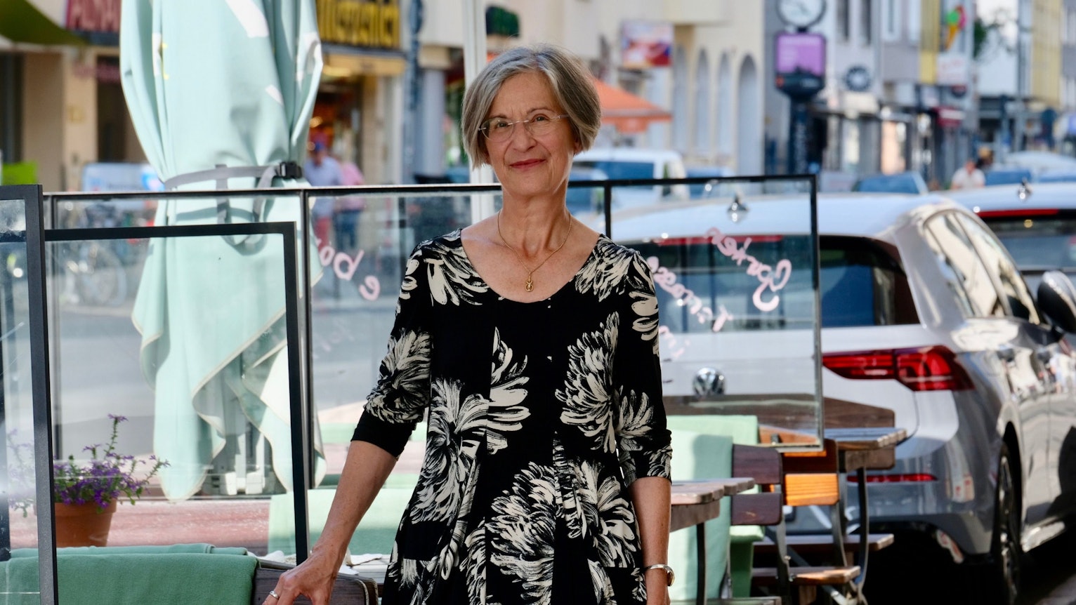 Eine Frau im schwarzen Kleid mit beigem Blumenprintmuster steht an einer Straße, an der viele Autos parken.