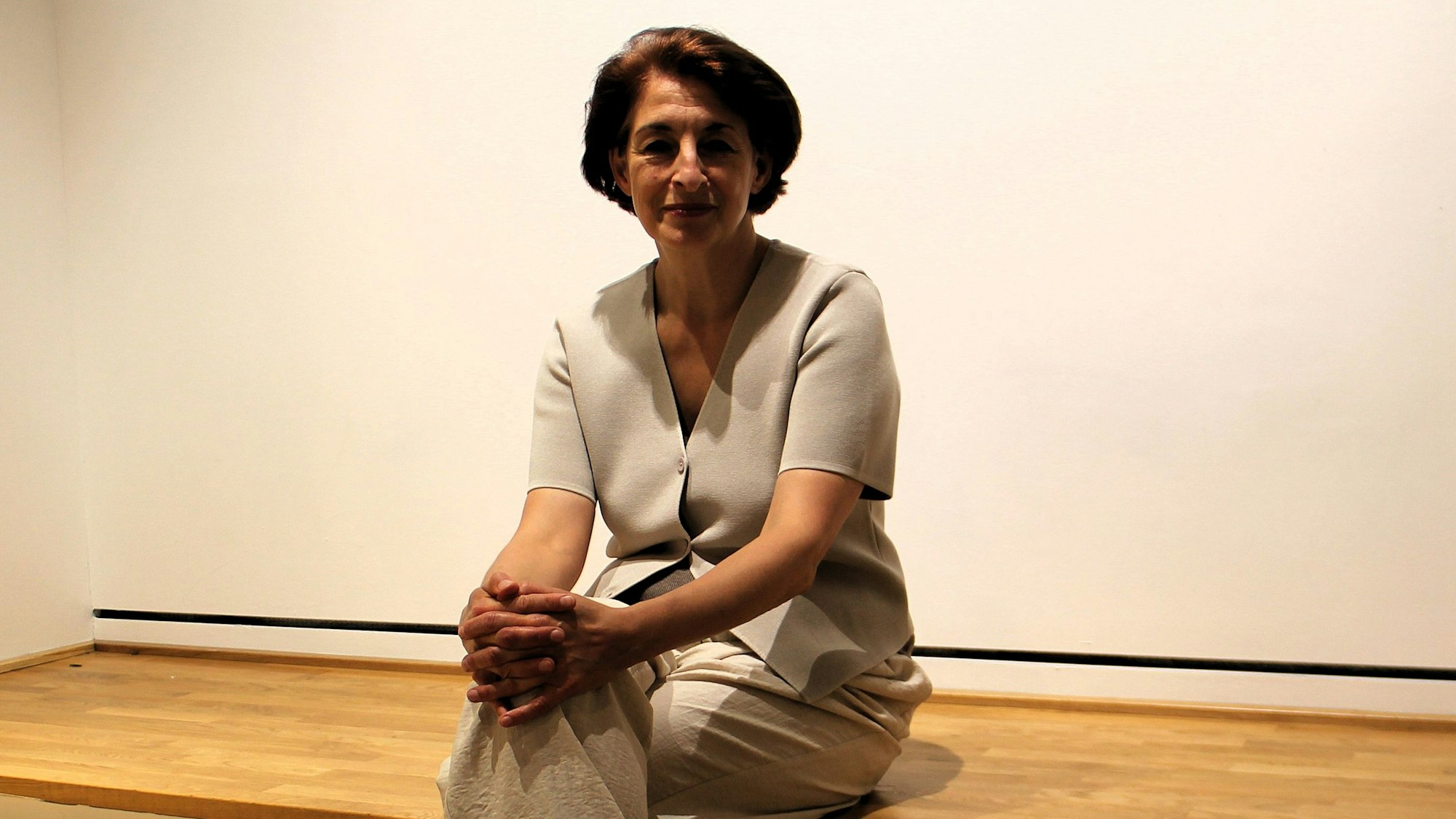 Eine Frau mit kinnlangen, dunkelbraunen Haaren trägt ein beiges Outfit und sitzt auf dem Rand einer Holzbühne.