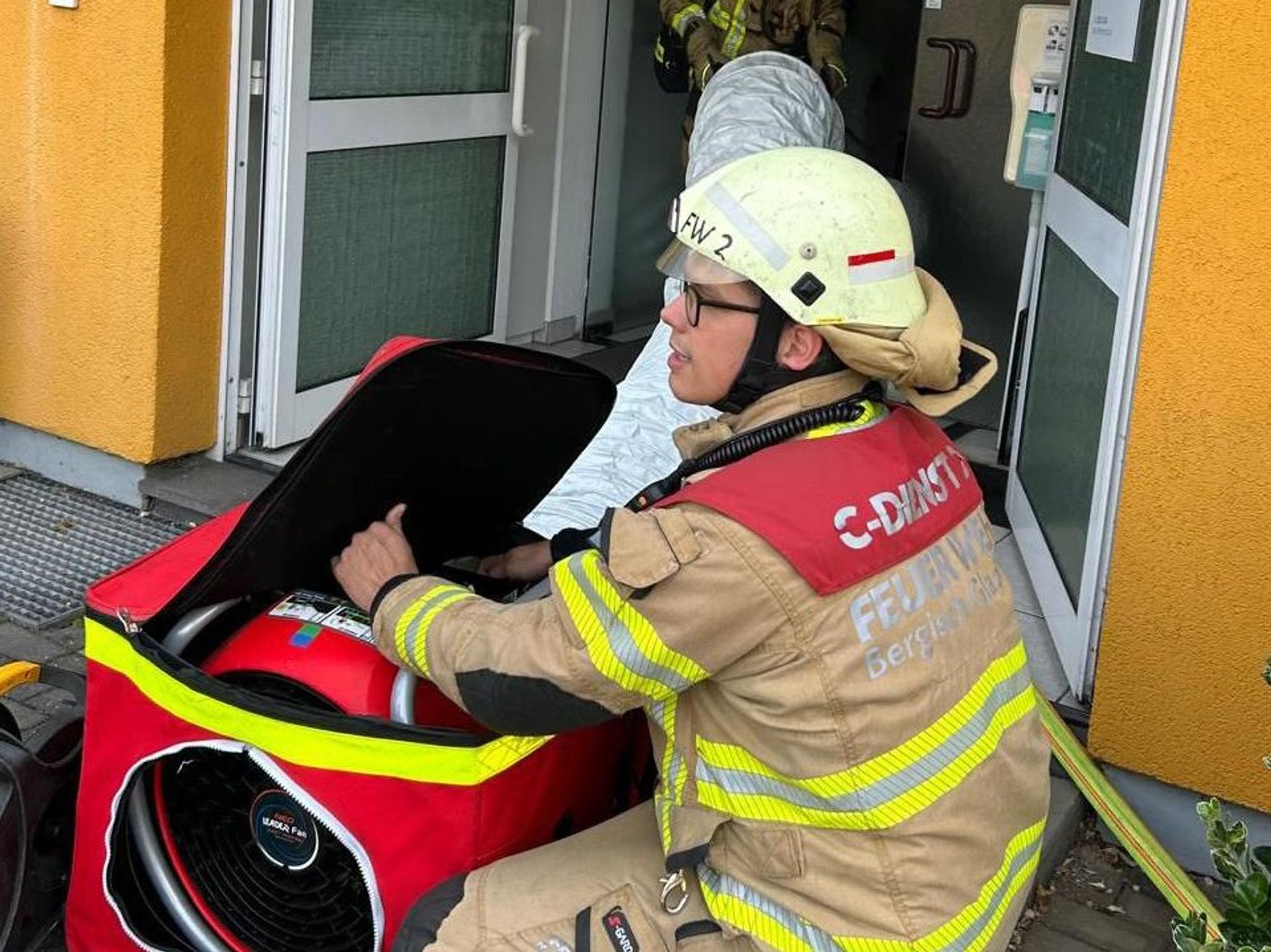 Feuerwehrleute stehen mit einem Gebläse an der Eingangstür eines Wohn- und Geschäftshauses an der Kürtener Straße in Bergisch Gladbach.