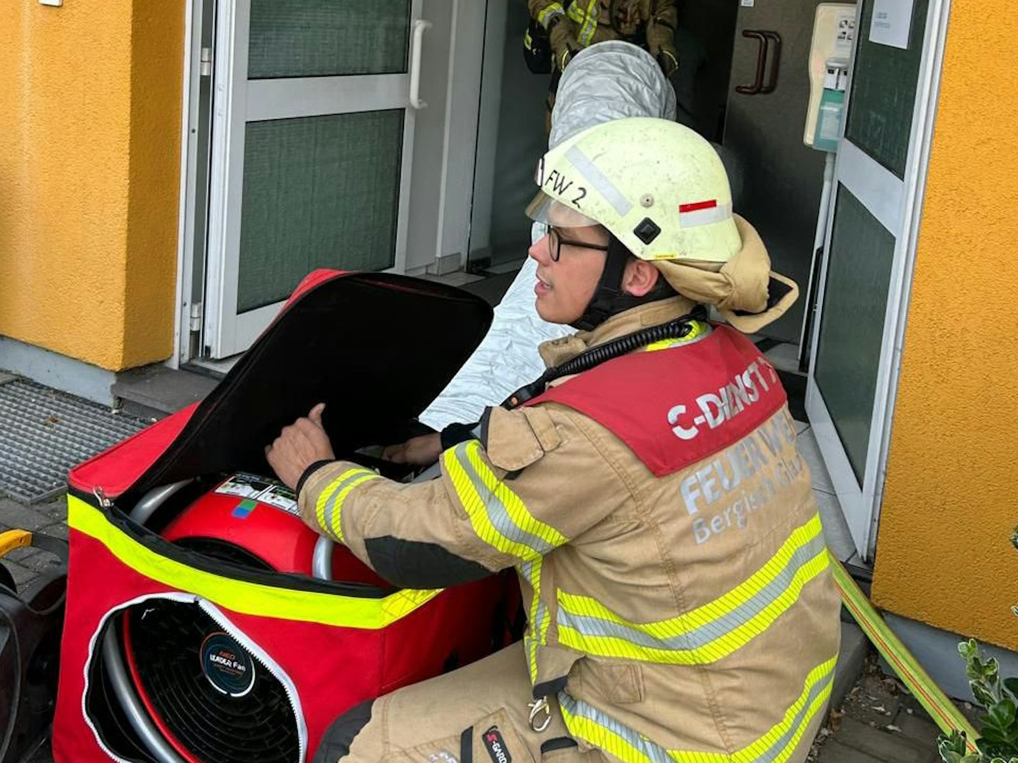 Feuerwehrleute stehen mit einem Gebläse an der Eingangstür eines Wohn- und Geschäftshauses an der Kürtener Straße in Bergisch Gladbach.