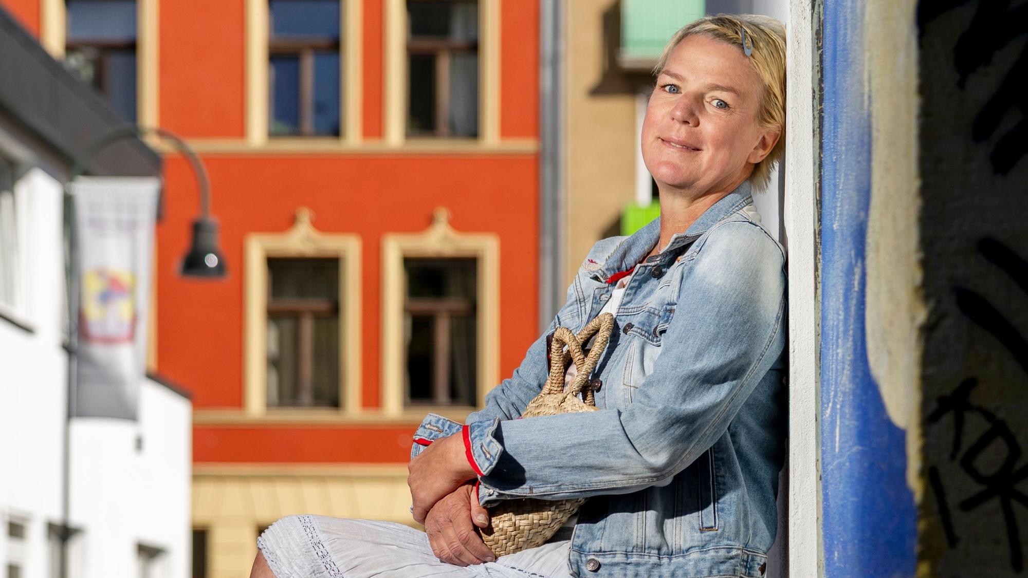 Eine Frau mit kurzen blonden Haaren und Minirock sitzt auf einem Steinblock vor einer Hauswand, im Hintergrund ein großes rotes Haus.