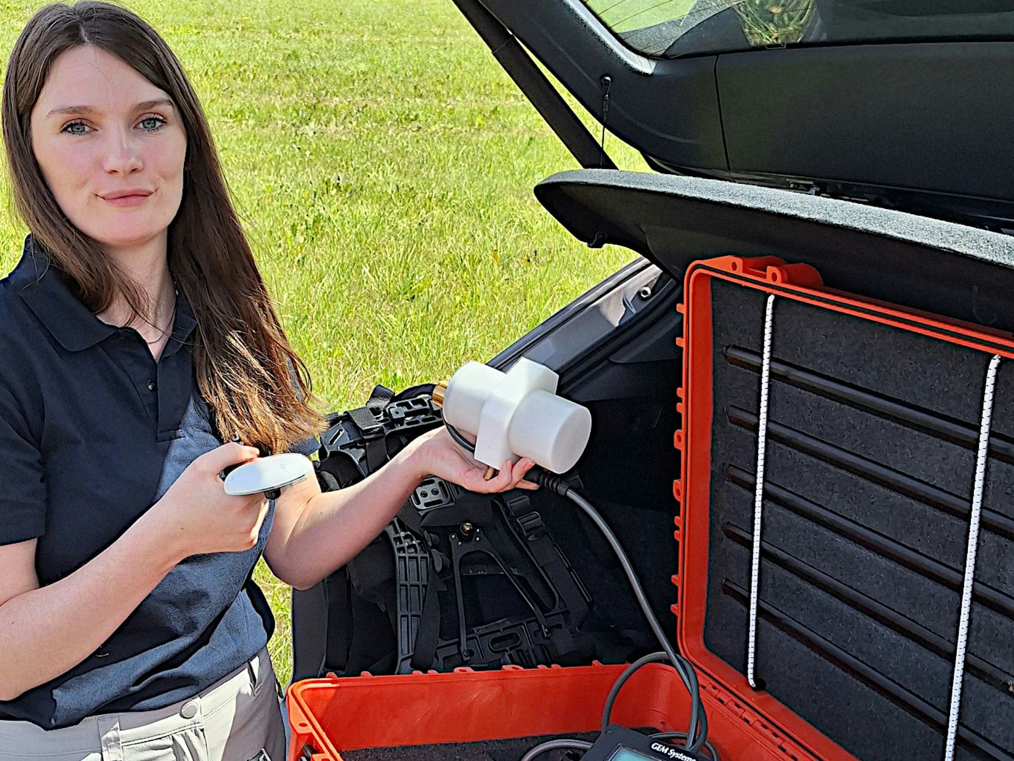Eine Frau steht an einem geöffneten Autokofferraum, darin eine Kiste mit wissenschaftlichen Apparaten.