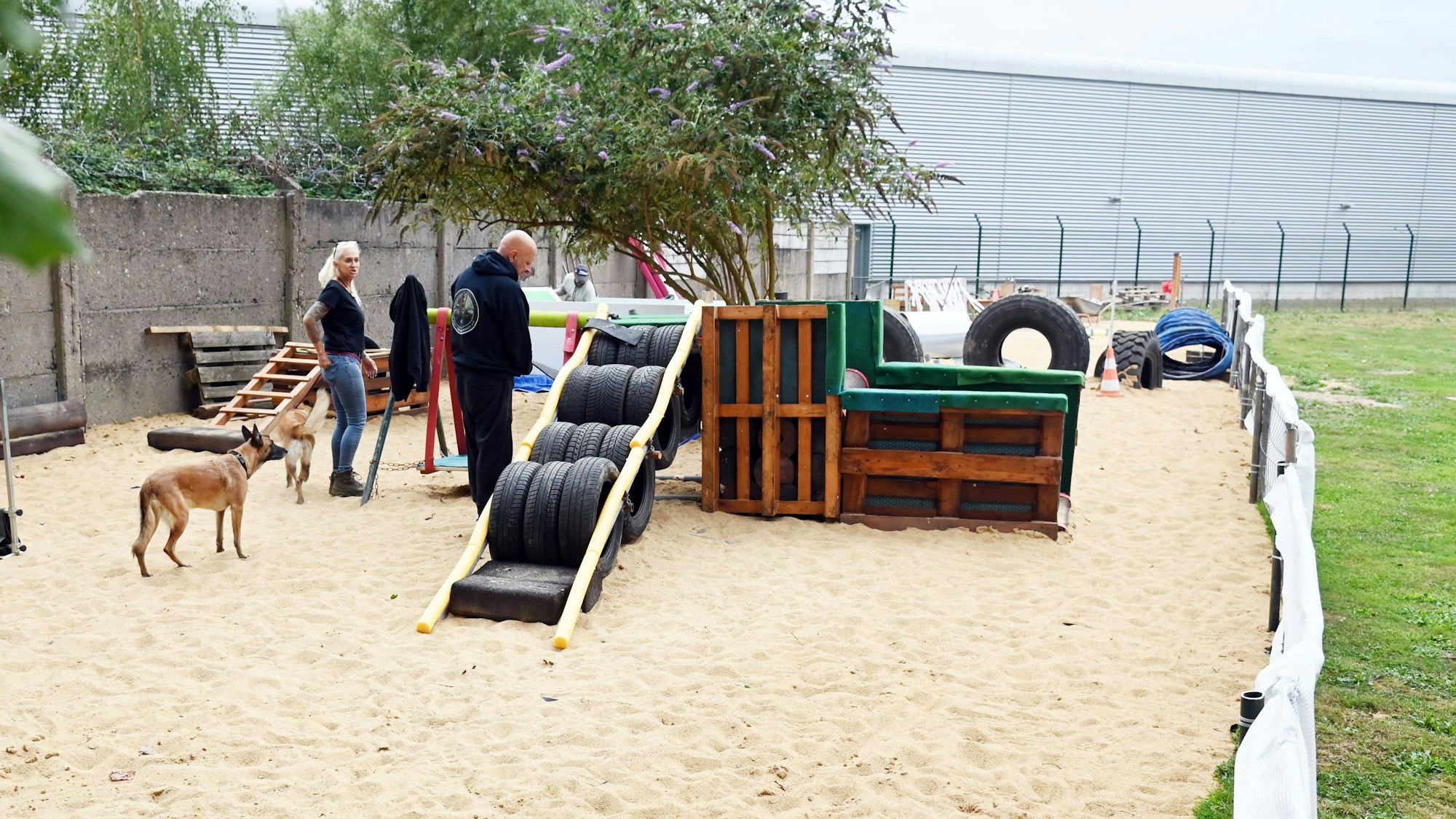 Auf einer Sandfläche mit einem Trainingsparcours stehen zwei Schäferhunde und zwei Menschen.