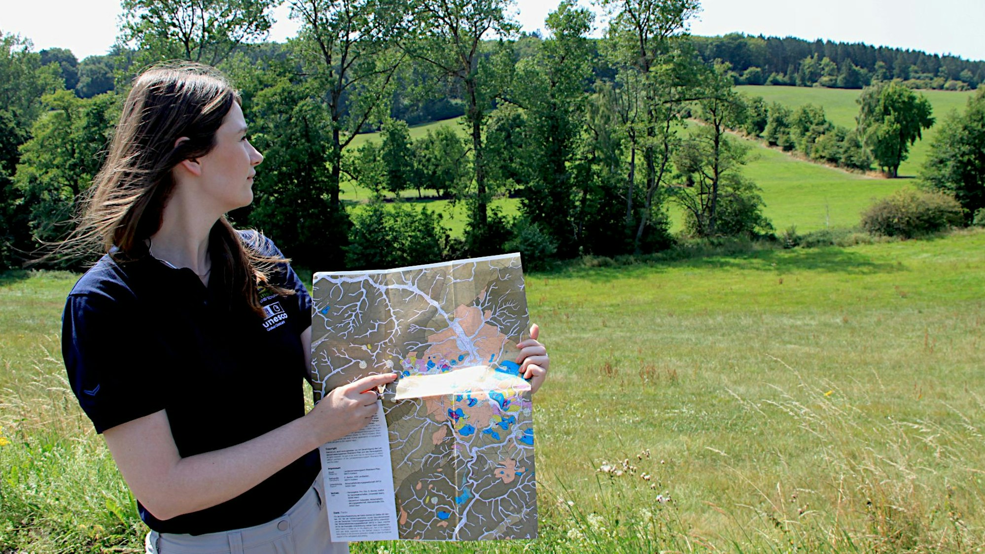 Eine Frau steht in einer Wiesen- und Baumlandschaft. Sie hält eine Landkarte in den Händen.
