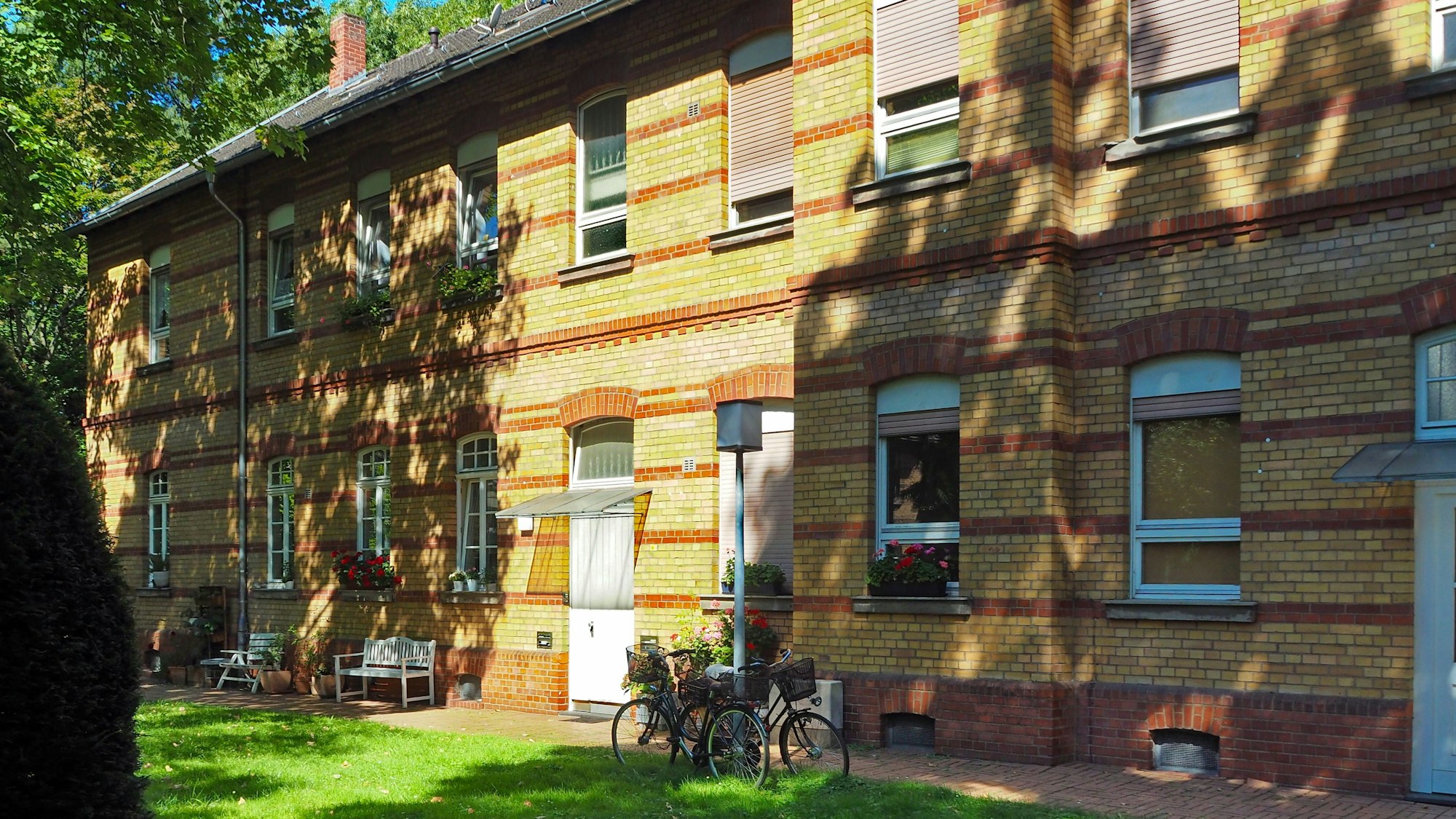 Vor zweigeschossigen Reihenhäusern stehen Bänke und Fahrräder.