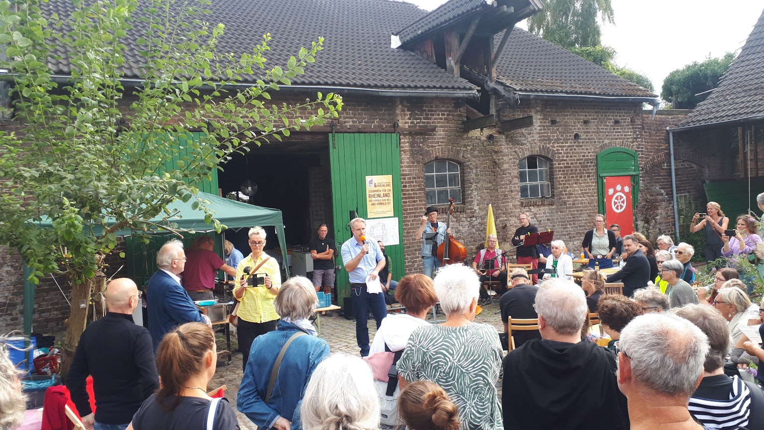 Ein Mann mit einem Mikro spricht vor zahlreichen Gästen. Im Hintergrund ist ein Hofgebäude zu sehen.