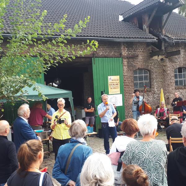 Ein Mann mit einem Mikro spricht vor zahlreichen Gästen. Im Hintergrund ist ein Hofgebäude zu sehen.
