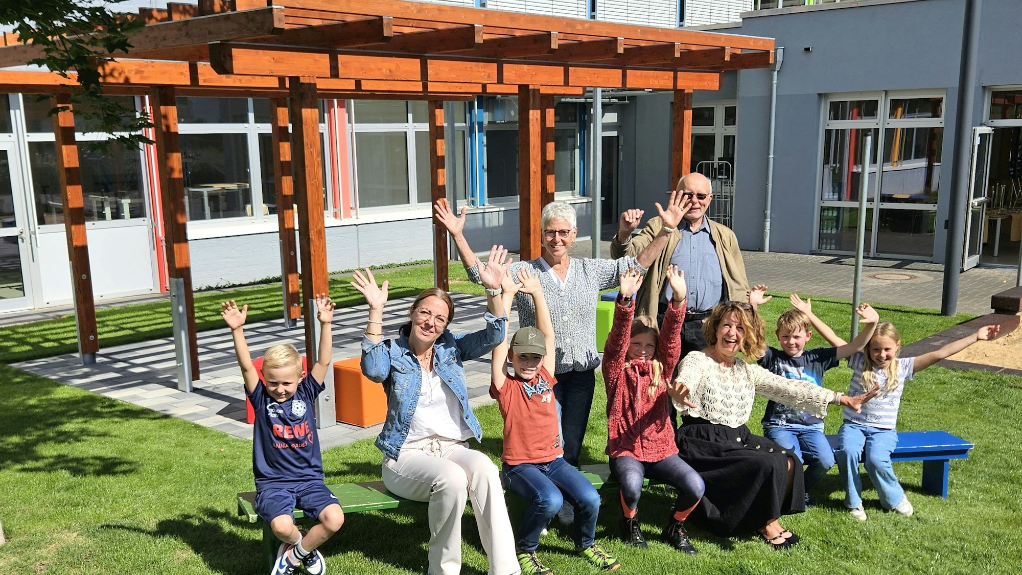 Kinder und Erwachsene sitzen auf Bänken vor der Pergola und winken mit den Händen.