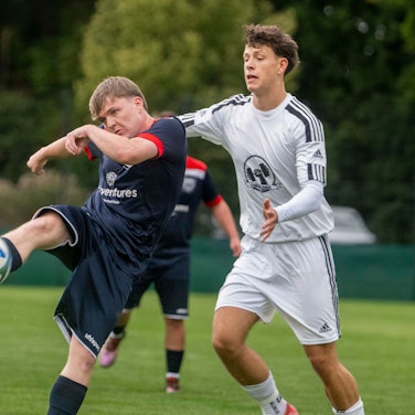 Ein Fußballer beim Torschuss, der Gegenspieler, der sich von hinten nähert, kommt zu spät.