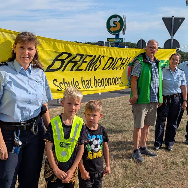 Vier Erwachsene und zwei Kinder stehen von einem Banner mit der Aufschrift „Brems Dich! Schule hat begonnen“.