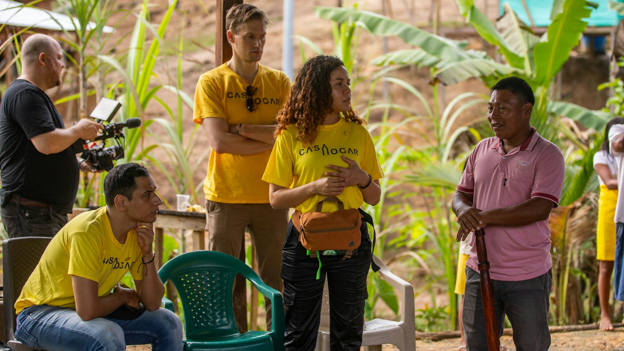 Menschen in gelben T-Shirts, auf denen der Casa-Hogar-Schriftzug steht, hören einem indigenen Mann zu. Im Hintergrund sieht man Palmen.