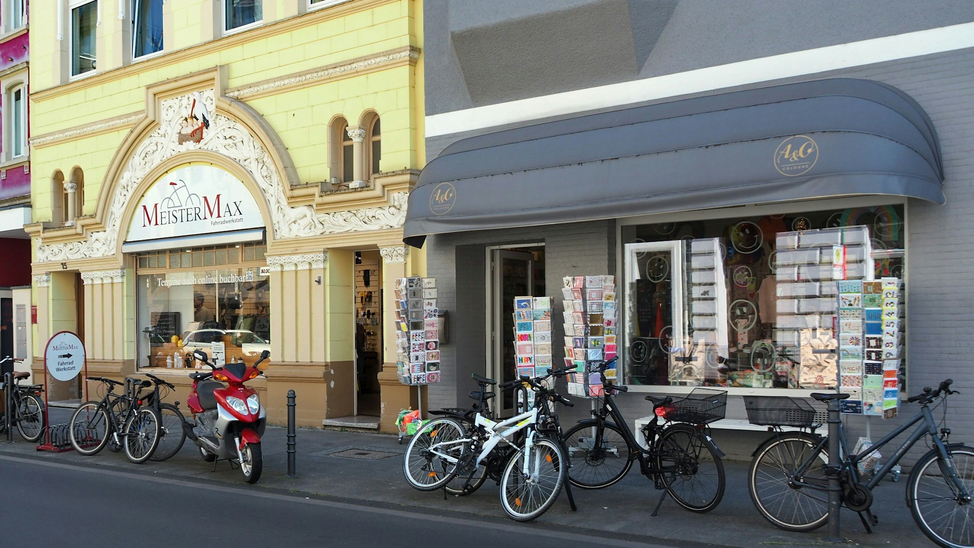 Zwei Häuser mit schönen Fassaden haben Geschäfte im Erdgeschoss, Fahrräder sind davor auf dem Bürgersteig abgestellt.