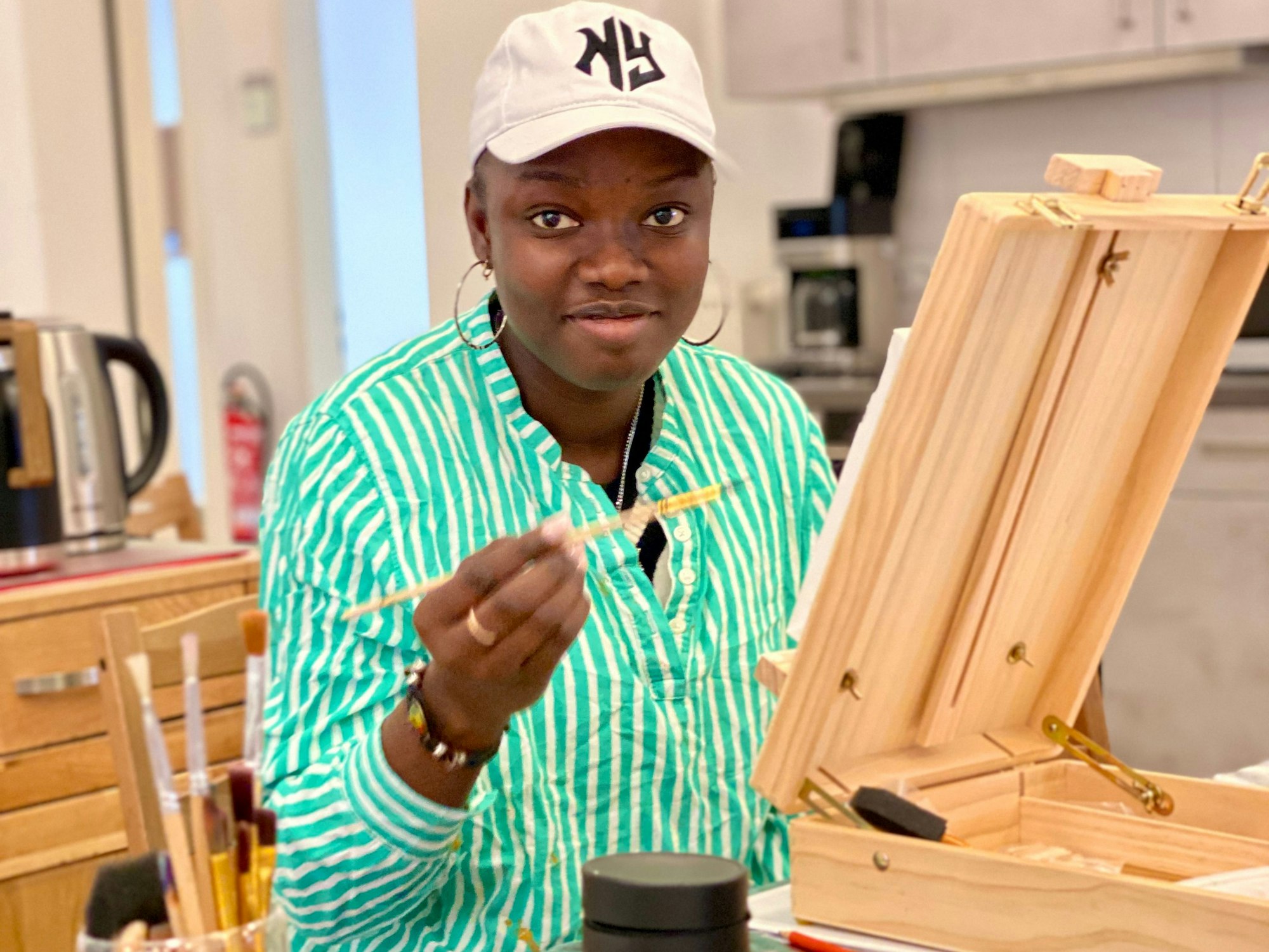 Eine junge Frau mit Baseballkappe sitzt vor einer kleinen Staffelei. In der Hand hält sie einen Pinsel.
