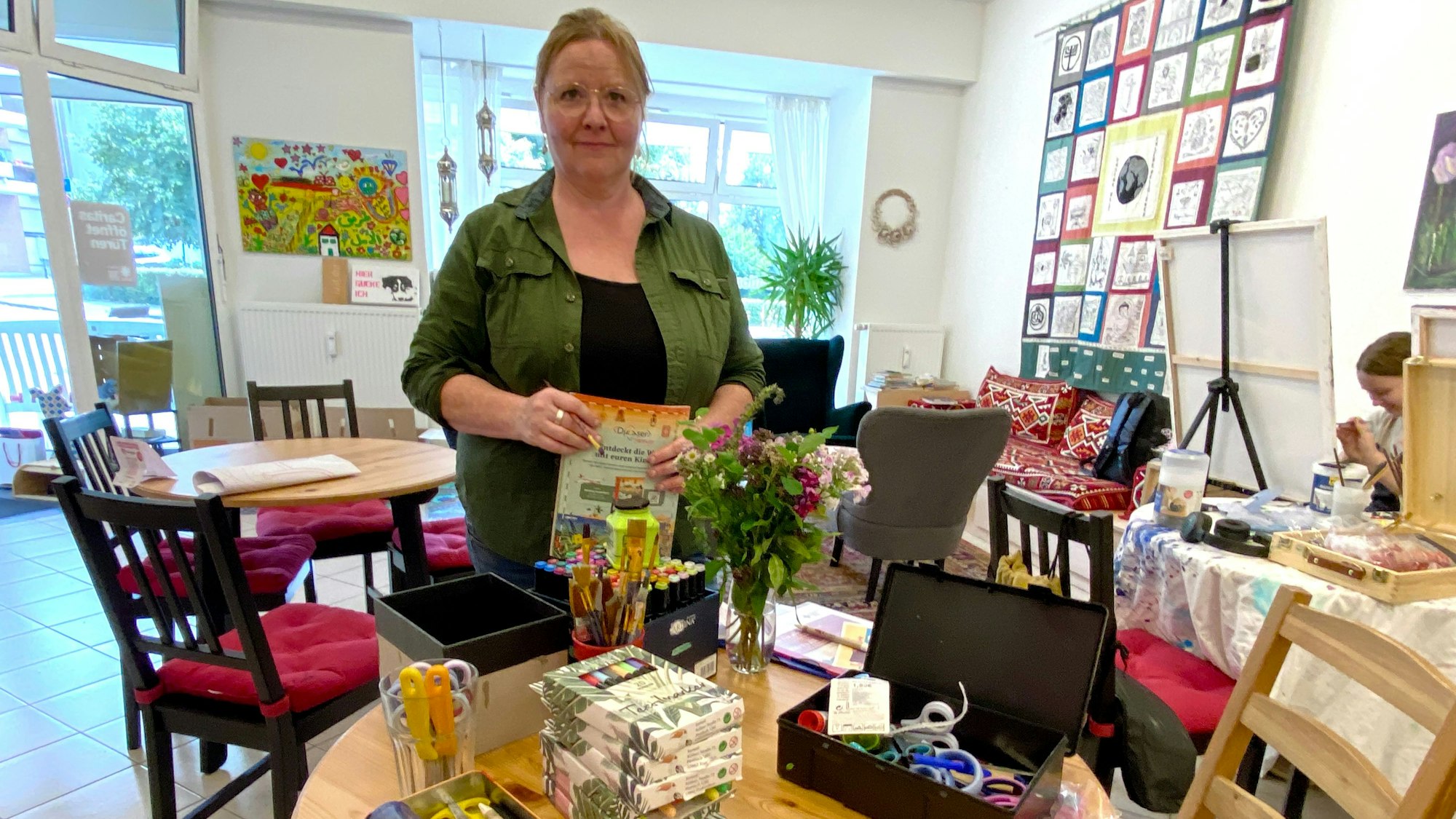 Eine Frau steht hinter einem Tisch, auf dem Farben, Stifte, Scheren und Bastelkram liegen.