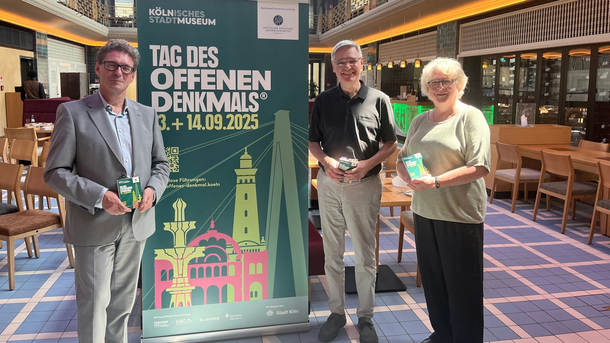 Drei Personen stehen vor einem großen Schild mit der Aufschrift „Tag des offenen Denkmals“ in einem alten Schwimmbecken, dass zu einem Restaurant umgebaut wurde.